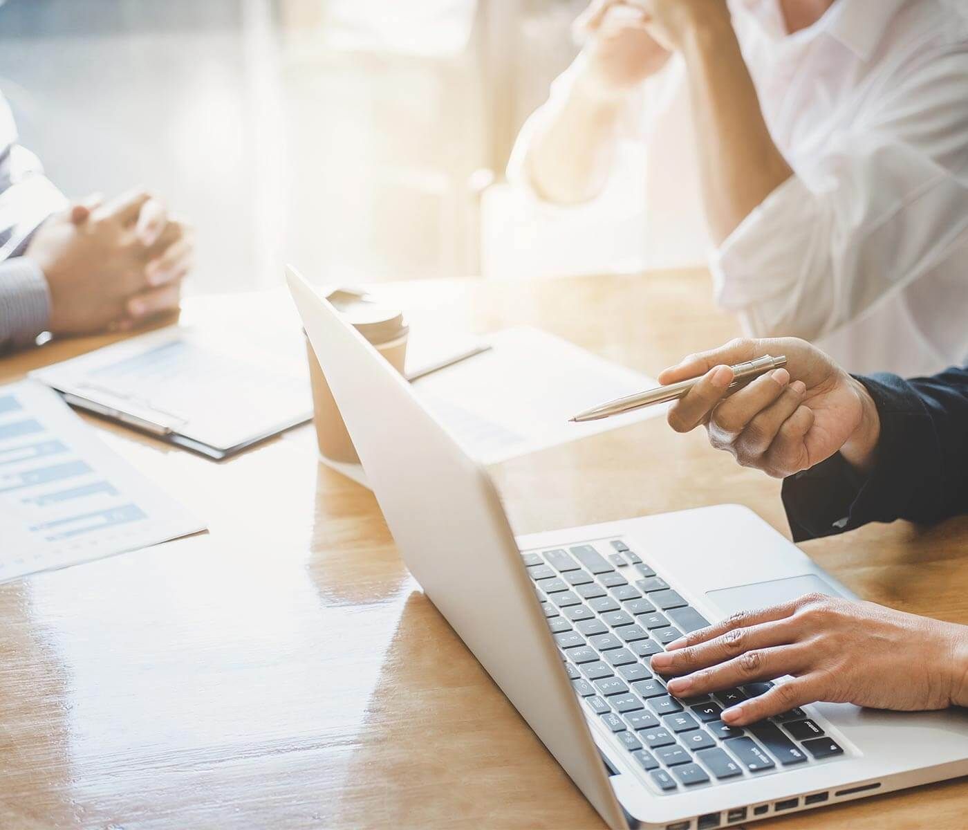 Sobre una mesa hay un ordenador portátil y sentadas a su alrededor hay varias personas, de las que solo se ve el torso. 